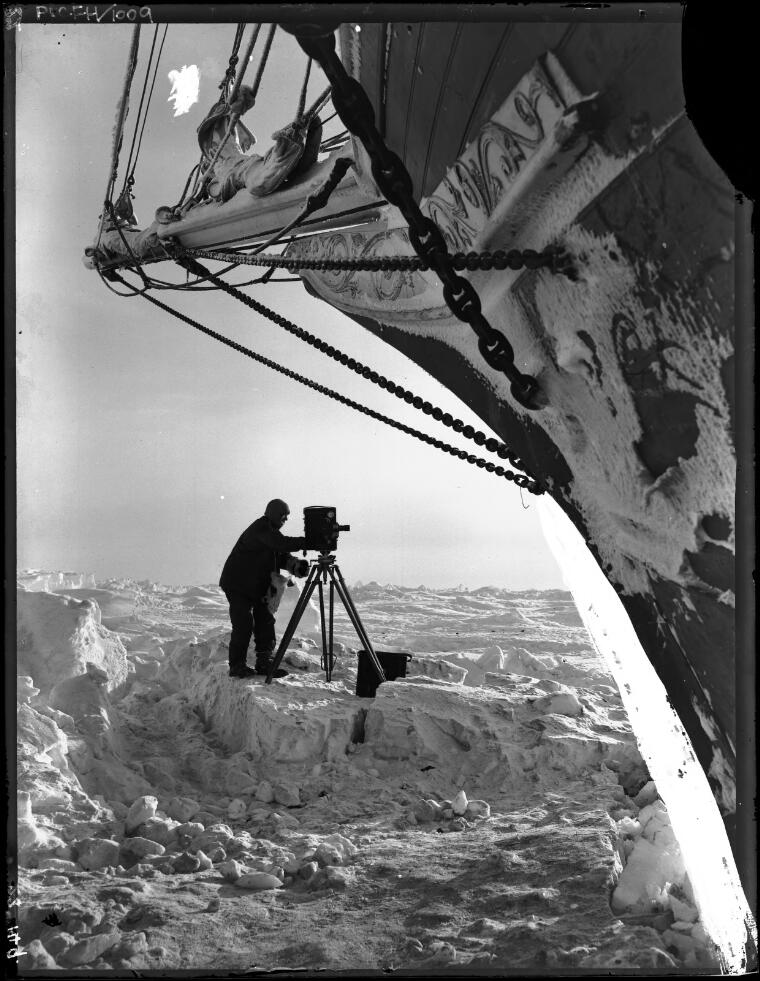 Frank Hurley with camera on ice in front of the bow of the trapped Endurance in the Weddell Sea, Shackleton expedition, 1915
