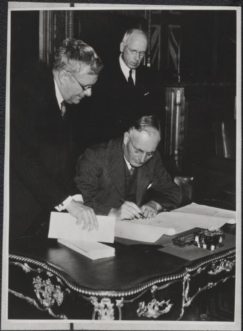 [Portrait of John Curtin seated at a desk with H. V. Evatt and Peter Fraser, Prime Minister of New Zealand, standing behind him, 1] [picture]