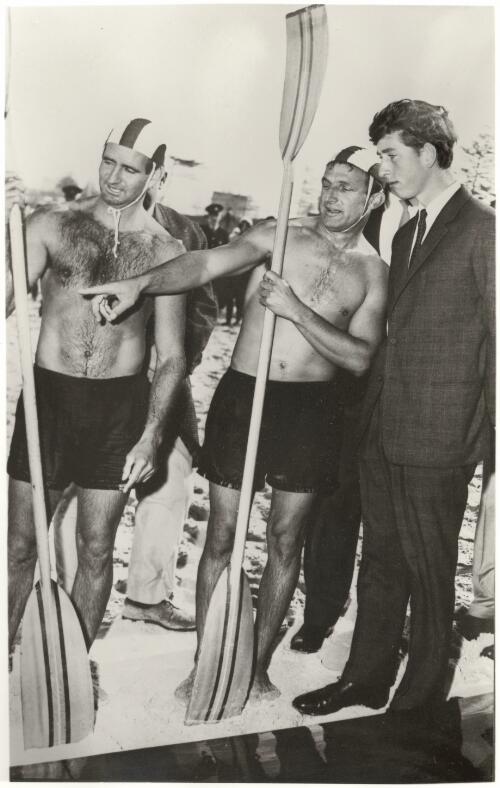 Prince Charles with Phil Coles and Ted Davis during the Prince's visit to North Bondi Surf Club, 25 May 1966, with Phil Coles explaining the details of a surf ski [picture] / Ern McQuillan