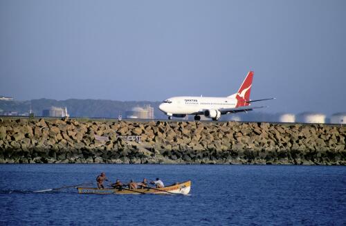 Lifesavers practice rowing on Botany Bay while a Qantas jet taxis along the north-south runway at Sydney (Kingsford Smith) Airport, New South Wales [picture] / Robert James Wallace