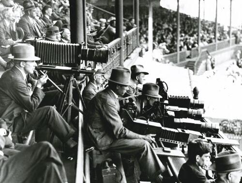 Photographers with folding cameras and spectators seated in a sports stadium, ca. 1930s [picture]