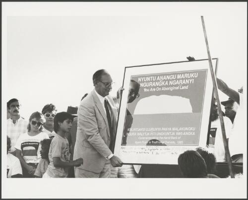 Sir Ninian Stephen holding a signed poster at the official ceremony of the handover of Uluru to its traditional owners, Northern Territory, 5 November 1985 [picture] / Department of Aboriginal Affairs