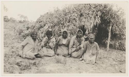 Five Aboriginal women sitting outside a gum tree branch shelter in missionary clothing, Northern Territory [picture]