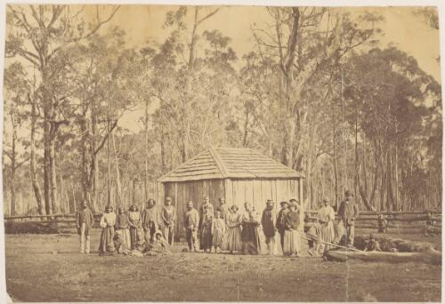 Group of Aboriginal people wearing European clothing outside a wooden hut with the forest behind them, a man in the centre wears a king plate, Warrnambool, Victoria [picture]
