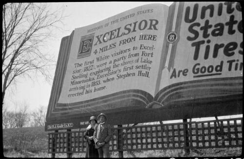 Billy Hughes and his wife Mary Hughes standing in front of a billboard, Excelsior, Lake Minnetonka, Minnesota [picture]