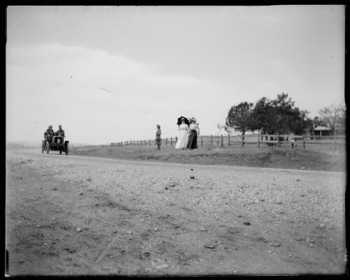 Men driving along the road in an early model car while two nurses look on, Gundagai, New South Wales [picture] / Charles Gabriel
