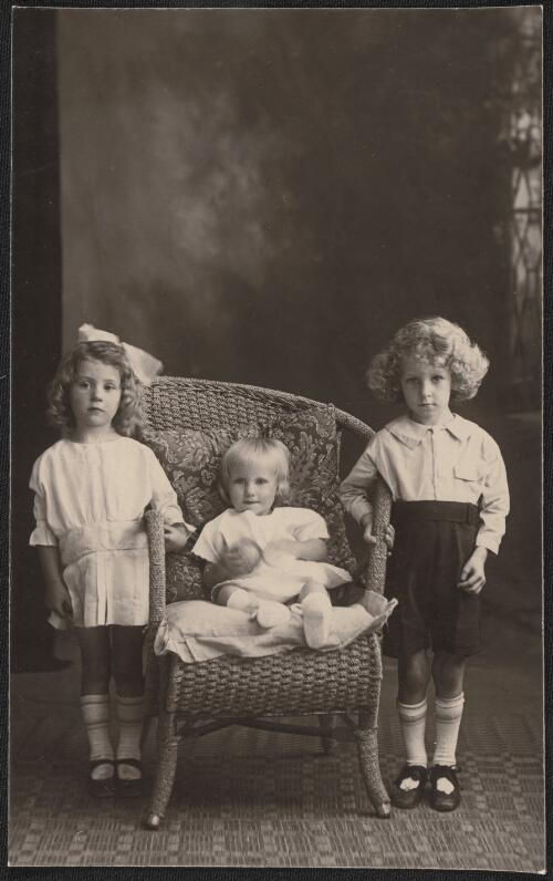 Two children standing and a smaller child sitting in a chair, Tumut, New South Wales  / R.C. Strangman