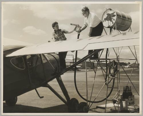 Australian Women Pilots' Association Air Reliability Trial entrant, Dr. Dorothy Rutherford is helped to fuel Fairchild 24 Argus II monoplane by an unidentified man on the tarmac at an airfield, 1953 [picture]
