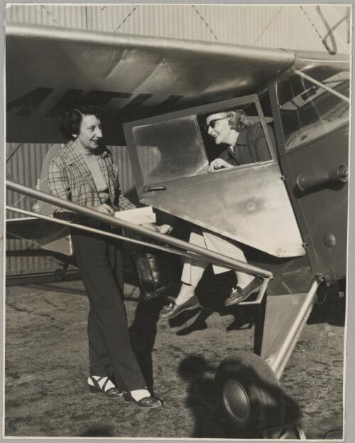 Dawn O'Mara helps Winifred Willmore step from the cockpit of an Auster J/4 Archer monoplane at an airfield, ca. 1953 [picture]