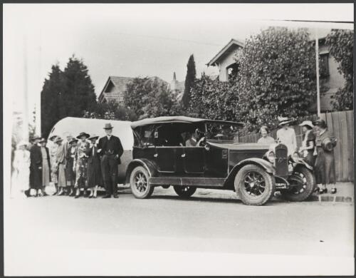 Group of people standing beside a car and caravan in a residential street, ca. 1925 [picture]