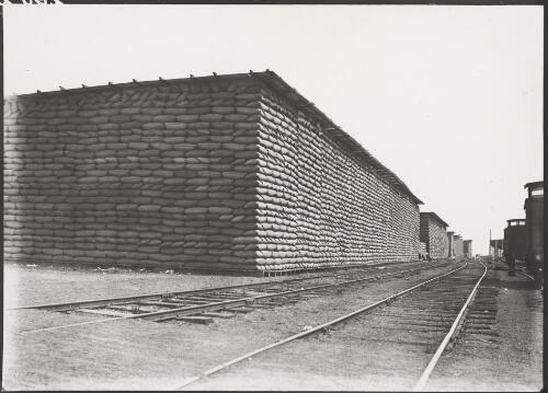 Stacked bags of wheat under a temporary corrugated iron roof at the railway siding, Sheep Hills, Victoria, ca. 1928 [picture]