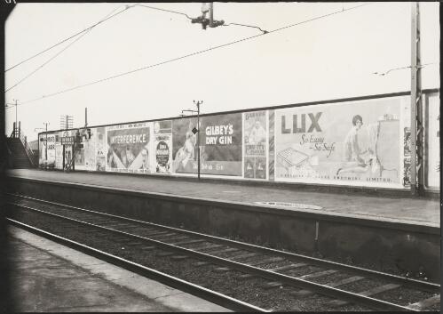 Advertising billboard for Foster's lager, Robur tea, Gilbey's dry gin, Barnes' fruity flakes, Lux soap flakes and New Majestic picture theatre screening of Interference on the railway platform, Burnley, Melbourne, ca. 1930 [picture]
