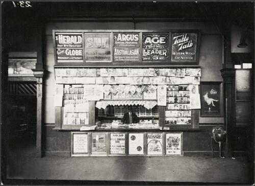 News stand in a railway station showing billboards publicising the opening of Parliament House in Canberra and an advertisement for Lustre Silktex stockings, Melbourne, 1927 [picture]