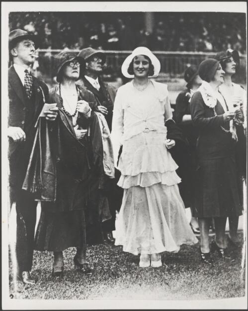 Young woman in a pale, bias-cut tiered dress standing with a group of men and women wearing dark clothes at the Melbourne Cup, Melbourne, 1931 [picture]