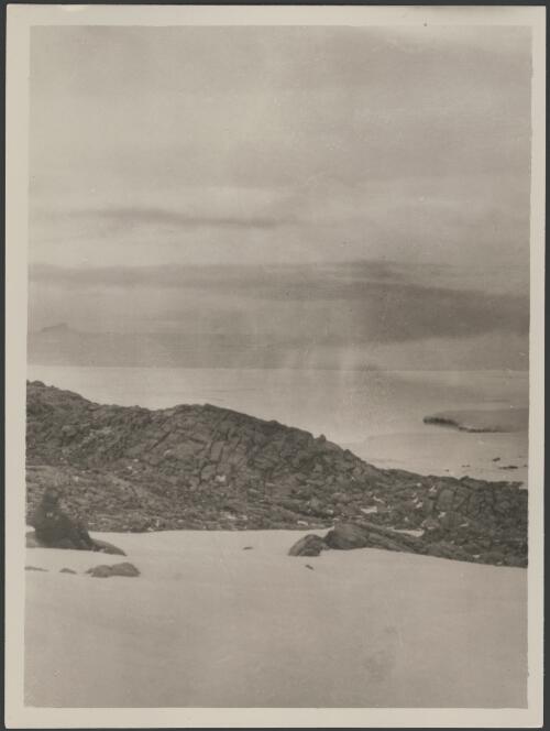 [View of Commonwealth Bay, explorer sitting on rocks in foreground, Australasian Antarctic Expedition, 1911-1914] [picture] / McLean