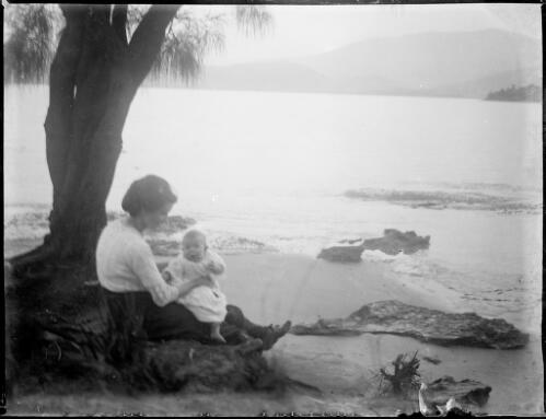 Woman holding a baby, seated under a tree beside a beach, Australia, ca. 1900 [picture] / E.W. Searle