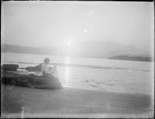 Woman holding a baby, seated on a rock beside a beach, Australia, ca. 1900 [picture] / E.W. Searle