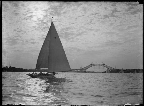 Sailing boat with the Sydney Harbour Bridge under construction in the background, Sydney Harbour, 1930 [picture] / E.W. Searle