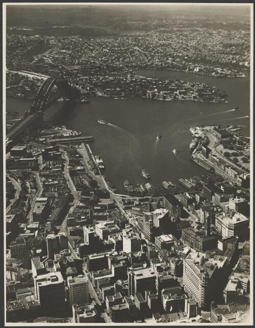 Aerial view of an incomplete Sydney Harbour Bridge with Circular Quay in the foreground, Sydney Harbour, 1931, 2 [picture] / E.W. Searle