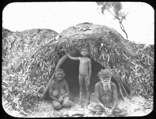 Aboriginal man and woman with a child outside wood and leaf shelter [transparency] / E.W. Searle
