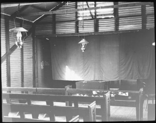 Hall interior with wooden pews, corrugated iron walls and gas lanterns [transparency] : miscellaneous glass slide / [John Flynn?]