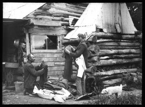 [Two unidentified men folding clothes outside a log cabin] [transparency] : a lantern slide used by John Flynn in lectures / [John Flynn]