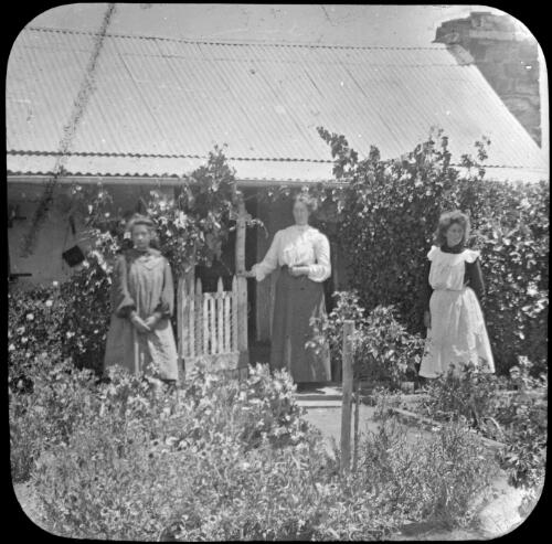 Three unidentified women standing outside an iron-roofed cottage, Angorichina Station, near Blinman, South Australia [transparency] : lantern slide used by Rev. F.H. Paterson, north South Australia / [John Flynn?]
