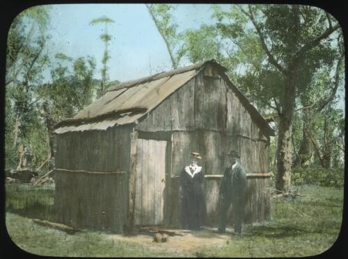 Two well dressed people standing outside a wooden hut [transparency] : a lantern slide used by John Flynn in lectures / [John Flynn]