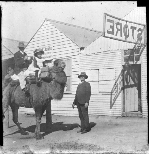 Three unidentified people, two on a camel, outside Bells and Jones Store, Blinman, South Australia [transparency] : lantern slide used by Rev. F.H. Paterson, north South Australia / [John Flynn?]