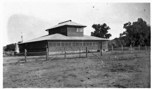 Wire fence in front of an unidentified building surrounded by foliage [picture] : Fred McKay Cape York Peninsula and West Queensland Patrol 1938-1940 / [Fred McKay]