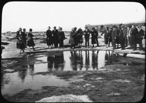 Group gathered beside large puddle of water in Coward Springs, train on railway in background [transparency] : a deputation slide of the AIM [Australian Inland Mission] Head Office, 1926-1940 / [John Flynn?]
