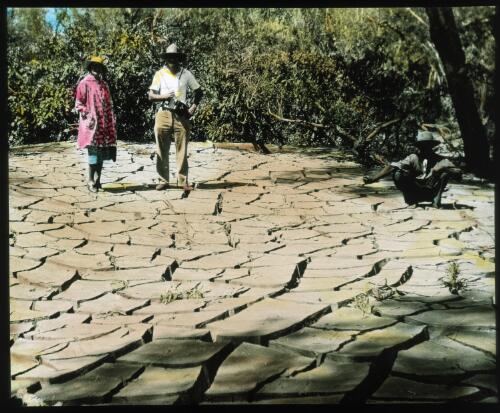Unidentified Indigenous man shows two unidentified people how the dry mud has cracked [transparency] : a lantern slide used by John Flynn in lectures / [John Flynn]