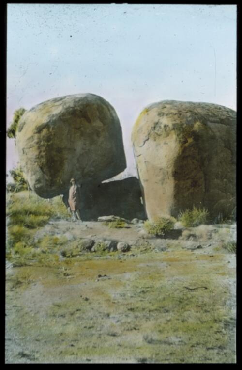 Unidentified woman standing between two large boulders [Devil's Marbles?] [transparency] : a lantern slide used for publicity in South Australia / [John Flynn]