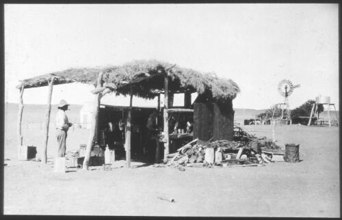Unidentifed man standing under thatched hut with windmill and water tank in distance, South Australia [transparency] : scene of mid-north South Australia used by Rev. F.H. Patterson on Stuart Patrol 1930+