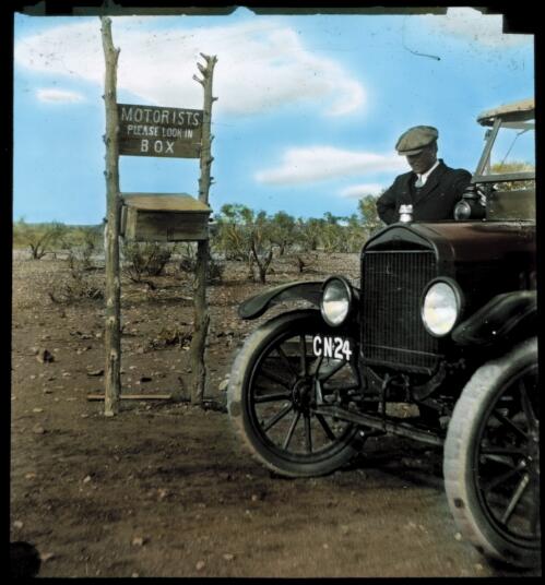 Unidentified man standing beside his car next to a mail box in the outback [transparency] : a lantern slide used for publicity in South Australia / [John Flynn]