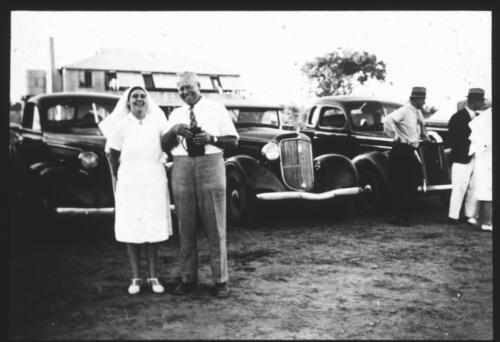 Portrait of Sister Banks and Mr Prior standing in front of a row of cars, Dunbar, Qld [transparency] : a lantern slide used for publicity in South Australia / [John Flynn]