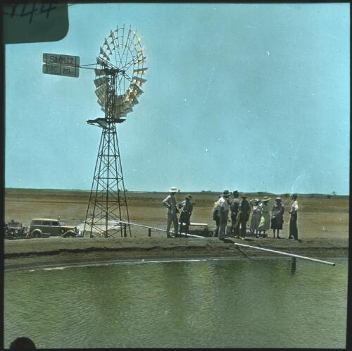 Group of people near a windmill and a dam, Birdsville Track [transparency] : a lantern slide used in lectures on all Australian Inland Mission activities, 1940- / [John Flynn?]