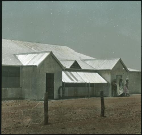 [Group of people standing next to a corrugated iron building] [transparency] : part of a lantern slide lecture collection / [John Flynn?]