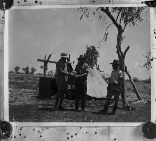 Two unidentified men holding unidentified objects at a campsite, with a third unidentified man watching [picture] : scenes of the Birdsville area / [John Flynn?]