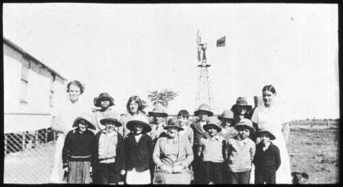 Portrait of three unidentified women and a group of children outisde a building with a windmill in the background [transparency] : a lantern slide used in lectures on all Australian Inland Mission activities, [1940-]/ [John Flynn?]