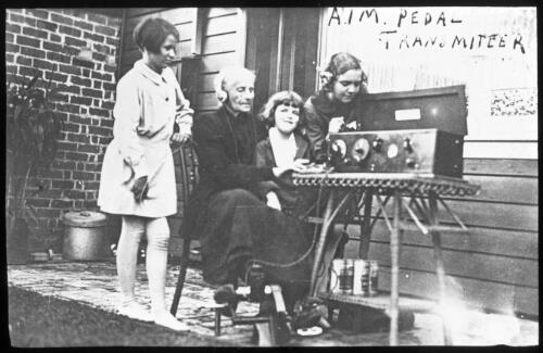 An elderly woman surrounded by three girls operating a pedal wireless on a porch, 1930 [transparency] / John Flynn