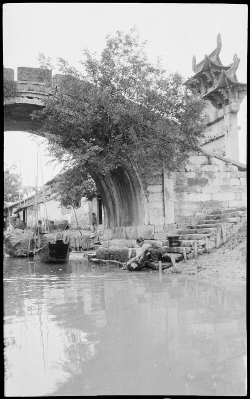 [Bridge on canal with large ornate gateway and stairway to canal where woman crouches, China, ca. 1930] [picture] / Stanley O. Gregory
