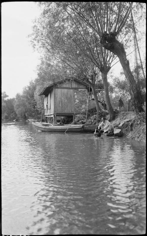 [Dwelling on canal bank with figure crouching nearby, China, ca. 1930] [picture] / Stanley O. Gregory