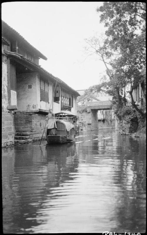 [Sampan with figure on the bow with pole alongside dwelling with bridge in the background, China, ca. 1930] [picture] / Stanley O. Gregory