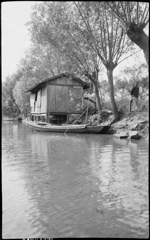 [Man standing on bank near dwelling on edge of water with a long empty punt alongside, China, ca. 1930] [picture] / Stanley O. Gregory