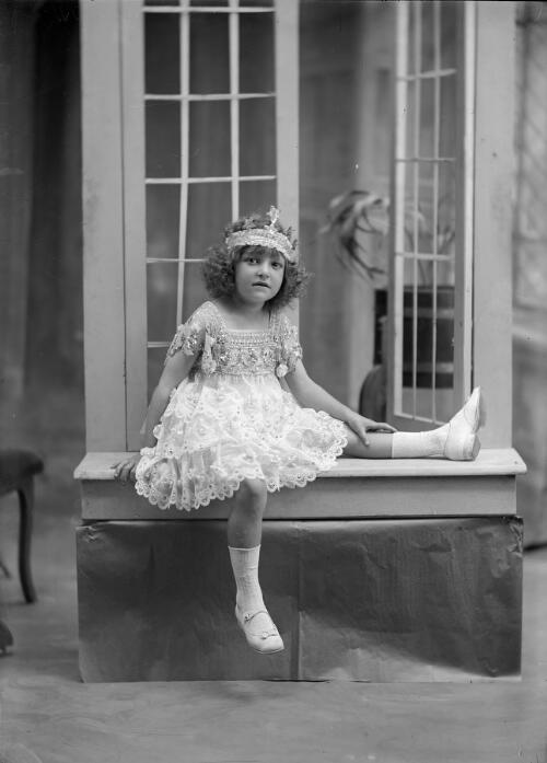 Child in ornate dress with bandeaux on head sitting on window seat in front of open window [picture] / Arthur William Emmerton