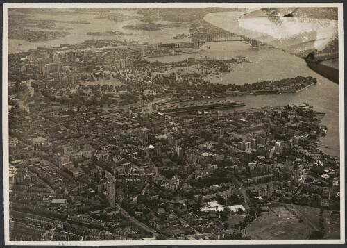 Aerial view of Sydney city from the south east, showing Sydney Harbour Bridge and part of aircraft wing [picture] / Sydney Morning Herald and Sydney Mail