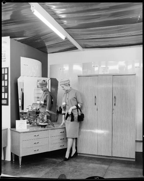 Unidentified woman standing next to a dressing table and cupboard in the Panelyte stand at the Royal Agricultural Showgrounds?, Sydney, 1 February, 1963 [picture] / John Mulligan