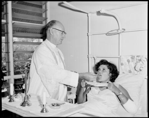 Father Frank Flynn serving communion to an unidentified Aboriginal patient in bed, Catholic Missions in the Northern Territory, 1 May 1962] [picture] / John Mulligan