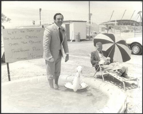 The Minister for the Capital Territory, Mr Hodgman, and Flipper, a plastic duck, pose in a wading pool erected by workman after Mr Hodgeman had turned the first sod at the site of the $7.8 million indoor swimming and training hall at the National Sports Centre, Bruce yesterday [picture] : the Commissioner of the NCDC, Mr Tony Powell, is at right / [photographed by Peter Wells?]